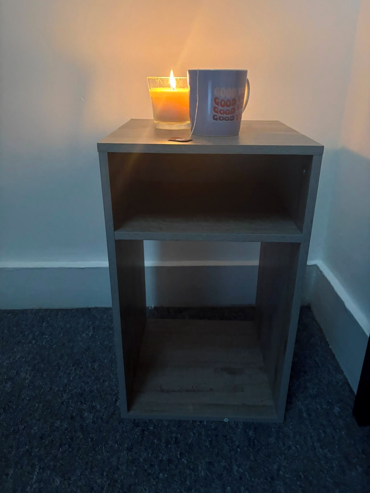 Un piccolo comodino in legno con un solo ripiano, una candela accesa e una tazza con la scritta 'GOOD MORNING'.