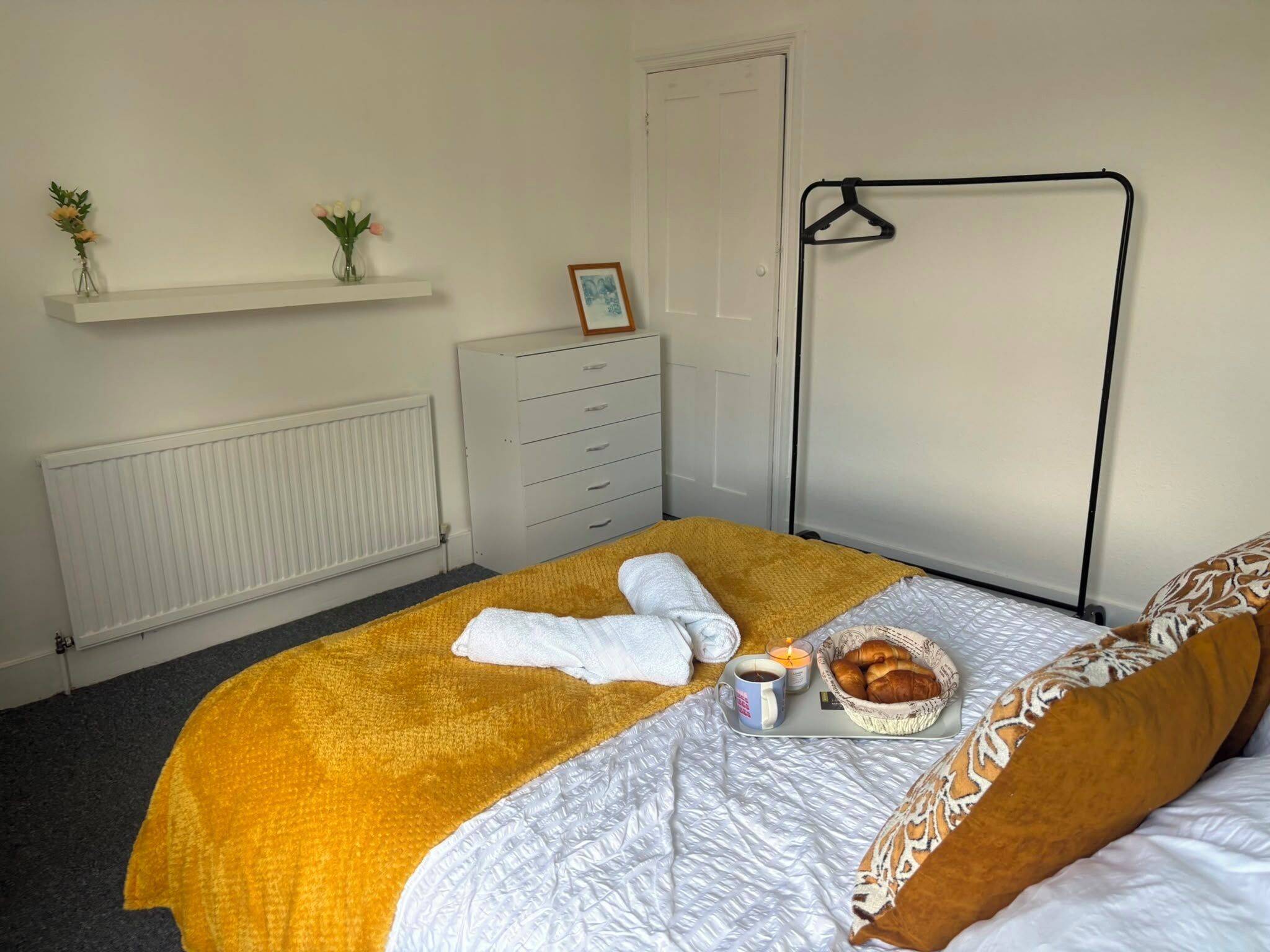 Een hotelkamer met een bed, een radiator, een dressoir, een kapstok en een plank met planten.