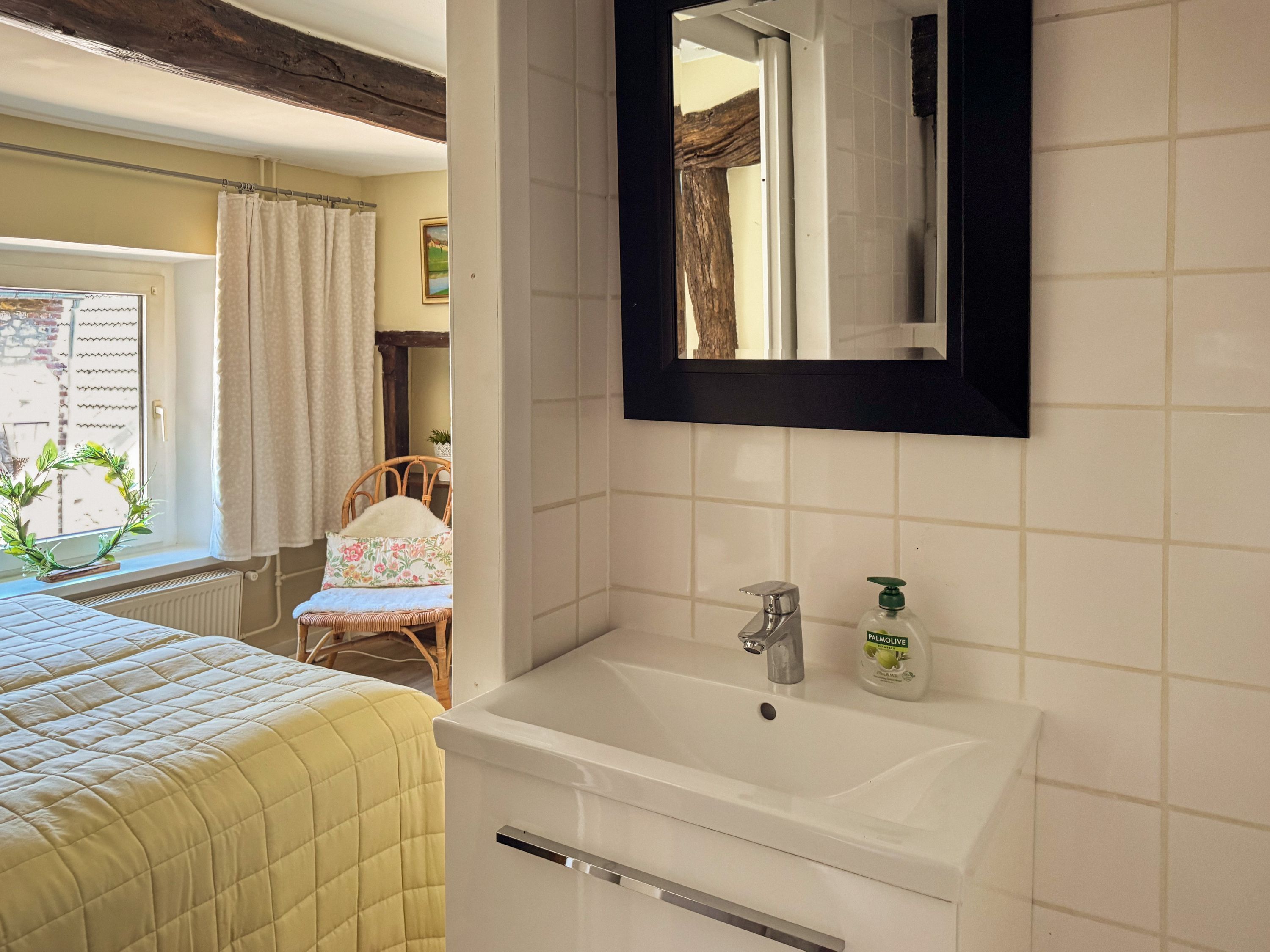 A bedroom with a bed, a window with curtains, a chair, a bedside table, and a bathroom with a sink and mirror.