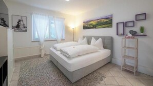 Un dormitorio con cama, ventana con cortinas, lámpara de noche, estante en la pared y un cuadro.