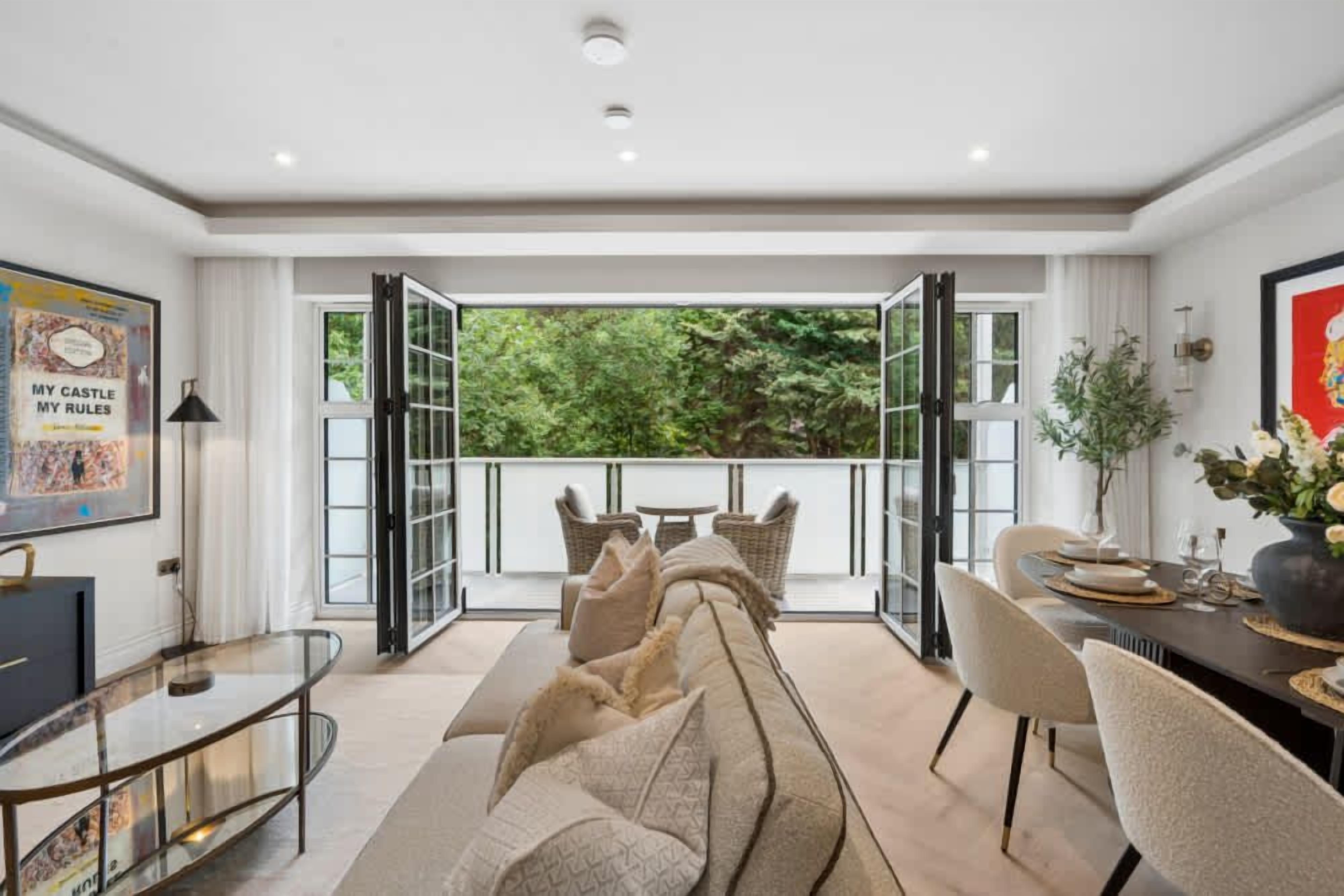 A modern living room with a sofa, dining table, and chairs, a balcony with glass doors, and a view of greenery.