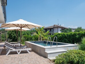 En swimmingpool med solstolar och parasoller, omgiven av grönska och ett hus i bakgrunden.