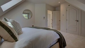 Een slaapkamer met een schuine kap, een ronde spiegel, een bed met kussens, en een deur die naar een andere kamer leidt.