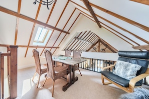 A loft bedroom with a sloped ceiling, wooden beams, a dining table with chairs, a sofa, and a view of the outside. - The Dairy (Corwen)