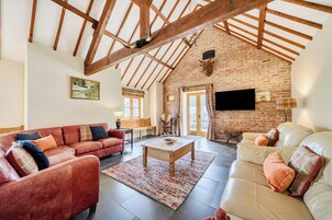 A spacious living room with a high ceiling, exposed wooden beams, a brick wall, a fireplace, and a television mounted on the wall.