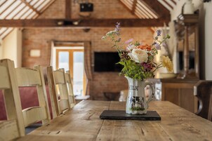 A rustic wooden table with a floral arrangement in a milk jug, set in a room with exposed brick walls and wooden beams.