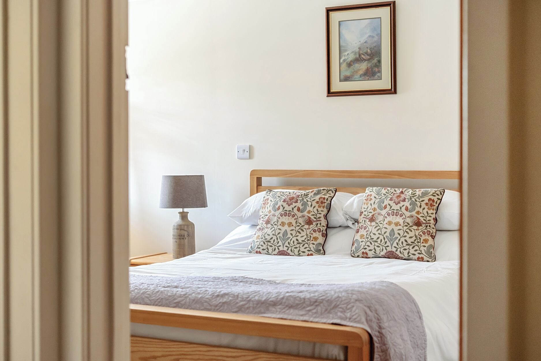 A bedroom with a wooden bed, floral pillows, a bedside lamp, and a framed picture on the wall.