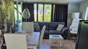 Una sala de estar moderna con un sofá, dos mesitas auxiliares, una mesa de comedor con sillas, una lámpara colgante y vistas a través de amplios ventanales.