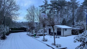 Zona residencial cubierta de nieve con una calle, un pequeño edificio blanco con puerta negra y árboles nevados.