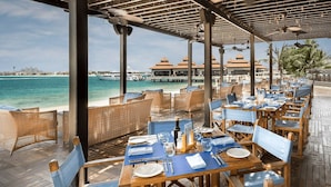Ein Restaurant am Meer mit Holztischen und -stühlen, Blick auf den Ozean und ein Gebäude mit rotem Dach im Hintergrund.