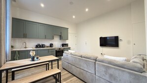 Eine moderne Küche mit grünen Schränken, einem Esstisch mit Bänken, einem grauen Sofa und einem an der Wand montierten Fernseher.