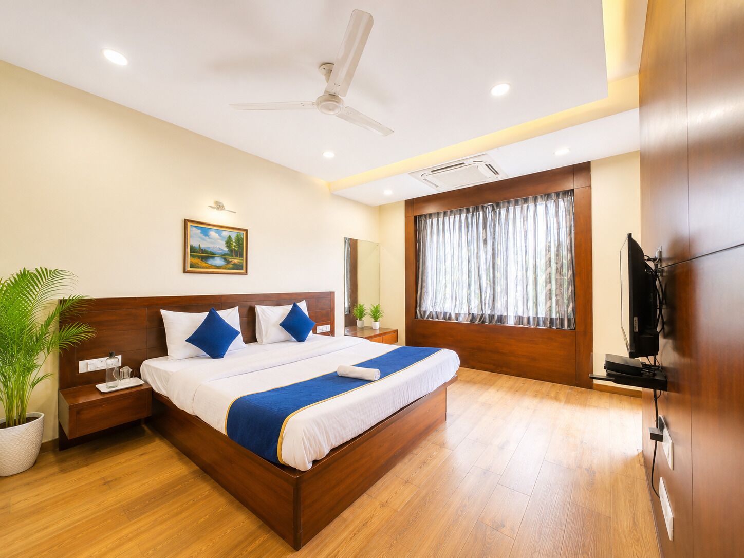 A modern hotel room with a wooden bed, a ceiling fan, a large window with curtains, a flat-screen TV, and a potted plant.