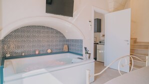 A modern bathroom with a glass-enclosed bathtub, a tiled backsplash, a mounted TV, and a visible kitchen area.