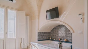 A room with a bed, a TV mounted on the wall, a bathtub, and a tiled backsplash.