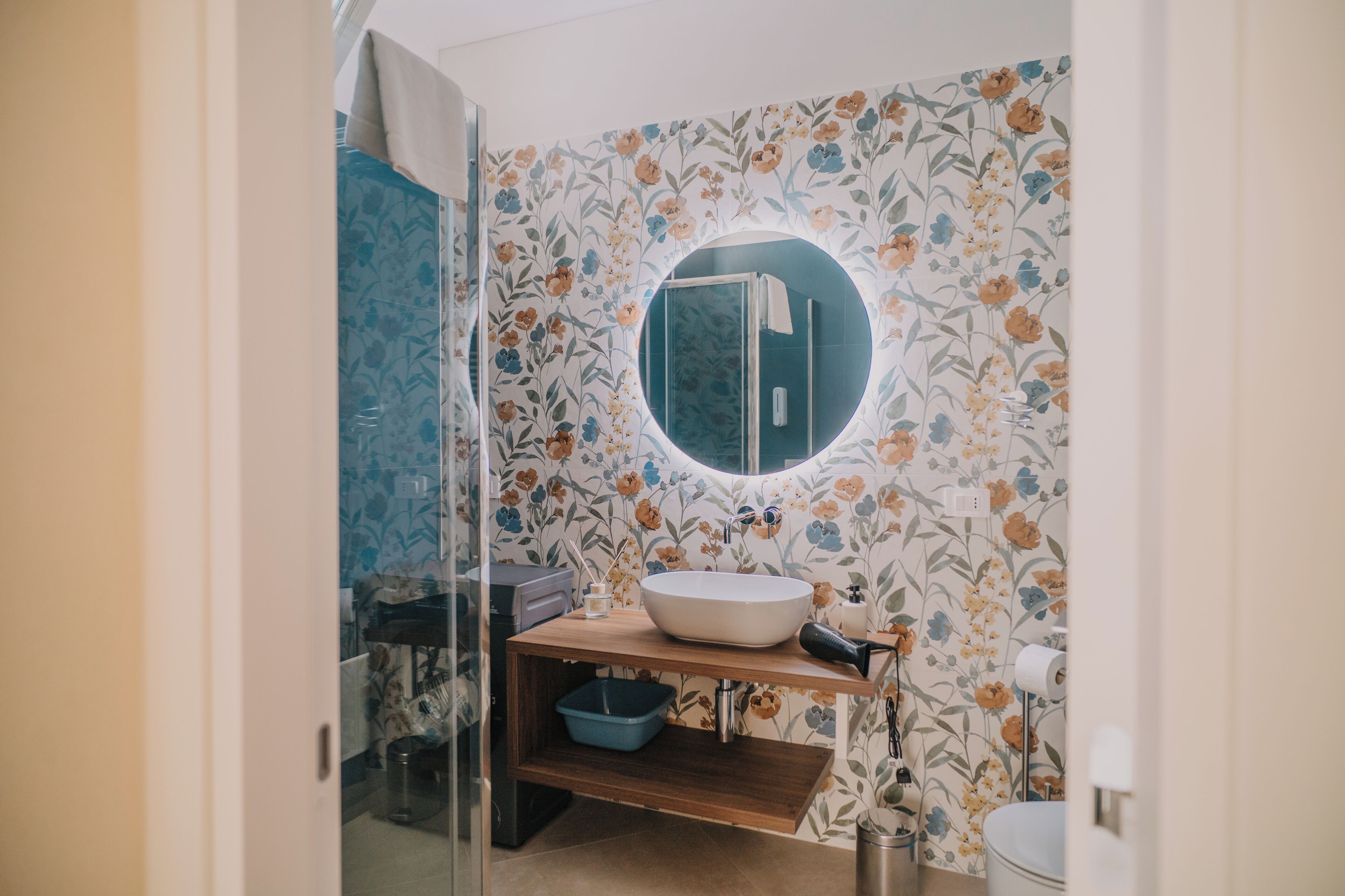 A bathroom with a round mirror, a wooden vanity, a blue basin, and floral wallpaper.