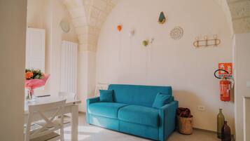 Une pièce avec un canapé de couleur turquoise, une table à manger blanche avec des chaises, une petite table sur laquelle est posé un vase de fleurs, et un extincteur fixé au mur.