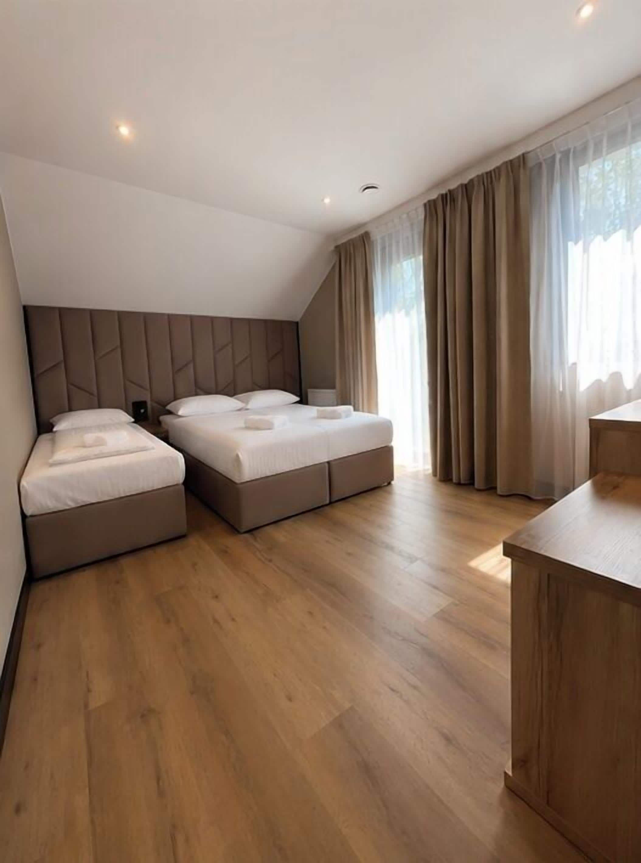 A modern hotel room with two beds, a wooden floor, a desk, and a large window with curtains.