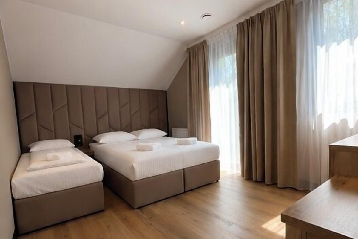 Moderni hotellihuone, jossa on kaksi sänkyä, puulattia, työpöytä ja suuri ikkuna, jossa on verhot.