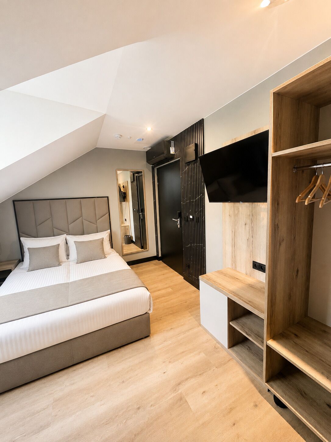 A modern bedroom with a sloped ceiling, a bed with a headboard, a wooden wardrobe, a television mounted on the wall, and a mirror.