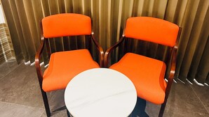 Duas cadeiras laranja com uma mesa redonda na frente, posicionadas diante de cortinas verticais.
