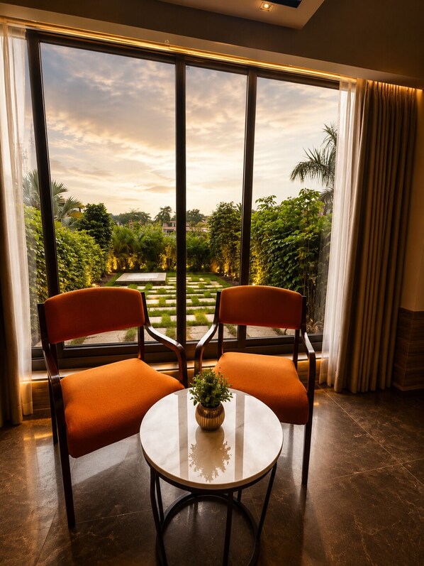 A room with two chairs and a table, large windows with a view of a garden, and a curtain on the right.