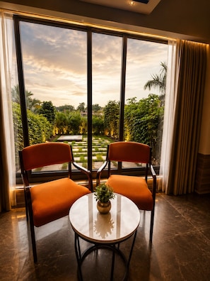 A room with two chairs and a table, large windows with a view of a garden, and a curtain on the right.
