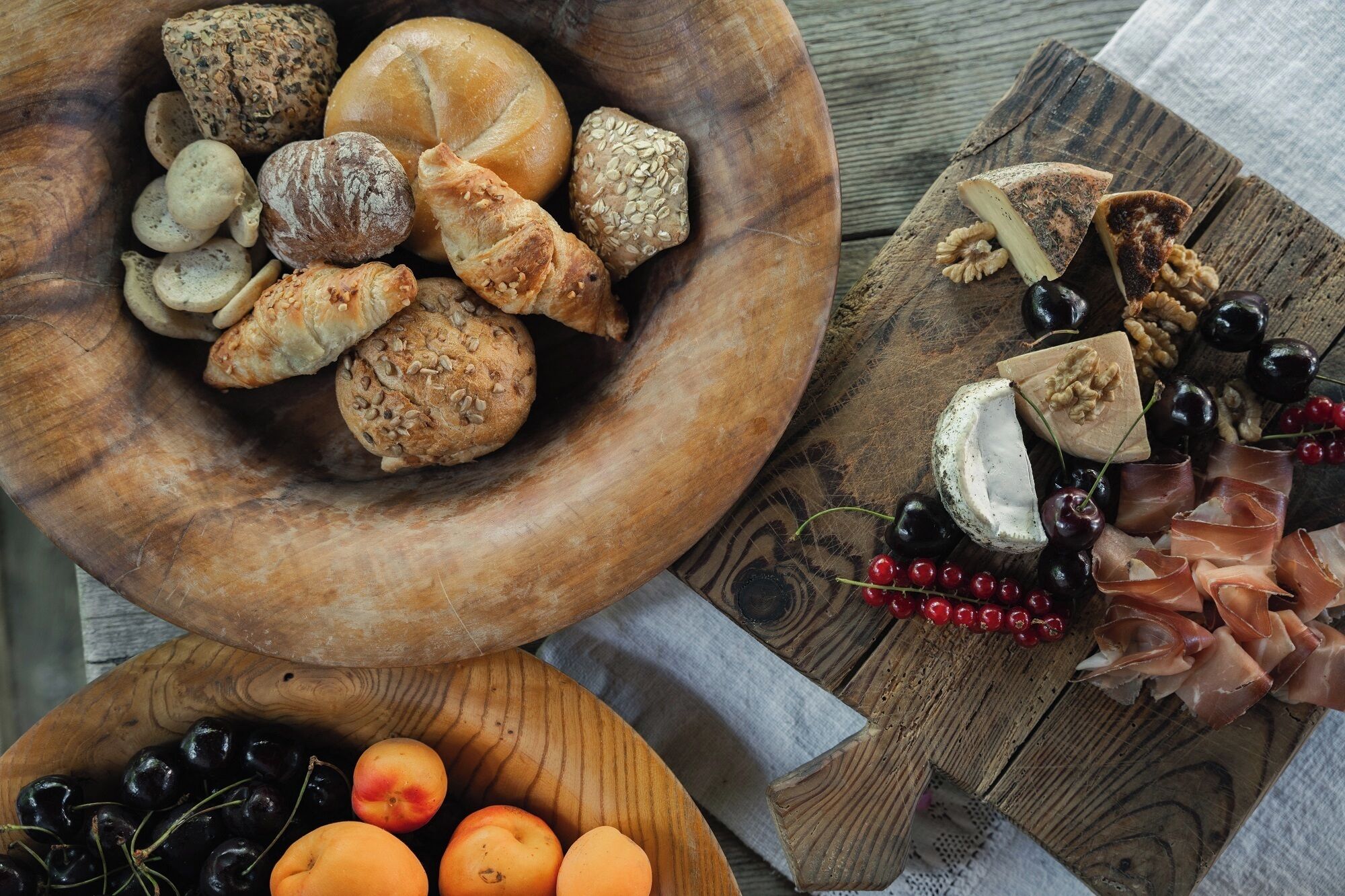 A wooden bowl filled with various breads and pastries, a wooden board with cheeses, nuts, and grapes, and a bowl of cherries and apricots.