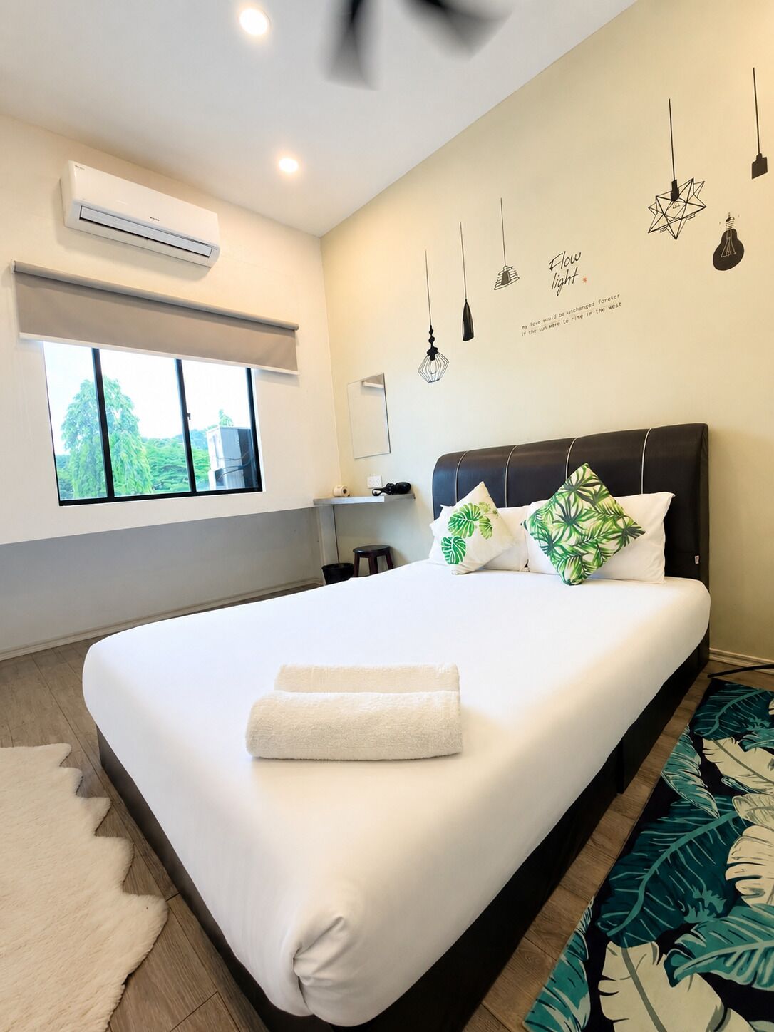 A modern hotel room with a large bed, an air conditioning unit, a window with a view, and a wall with hanging lights and a motivational quote.