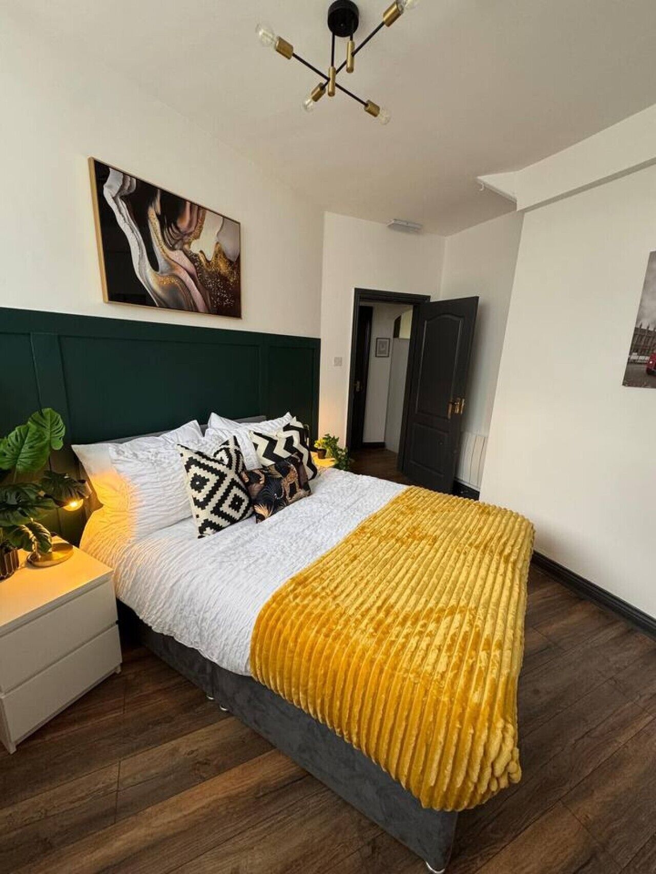 A bedroom with a bed, a nightstand, a plant, a painting, and a door.
