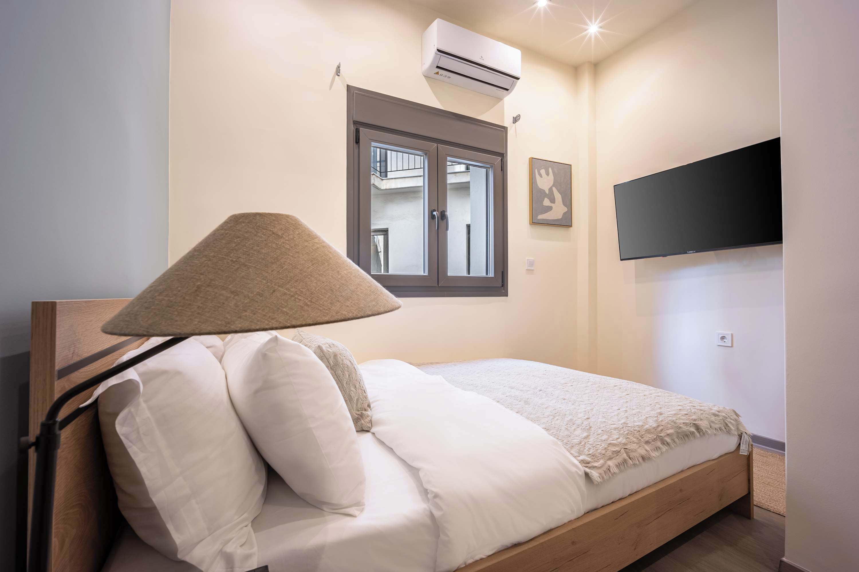 A bedroom with a bed, a lamp, an air conditioner, a window, and a TV mounted on the wall.
