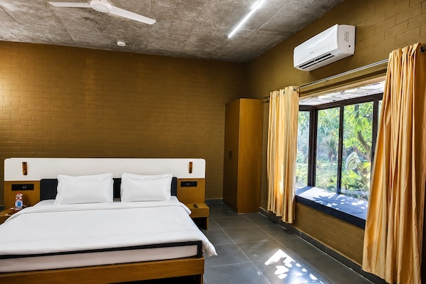 A hotel room with a bed, a nightstand, an air conditioning unit, a window with curtains, and a view of greenery outside.
