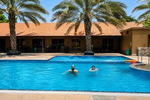 A swimming pool with two people in the water, surrounded by palm trees and a building with a tiled roof.