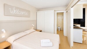 Et hotelværelse med en seng, et sengebord, en væglampe, et skilt med teksten 'Welcome', et badeværelse, der kan ses igennem en åben dør, og et tekøkken.