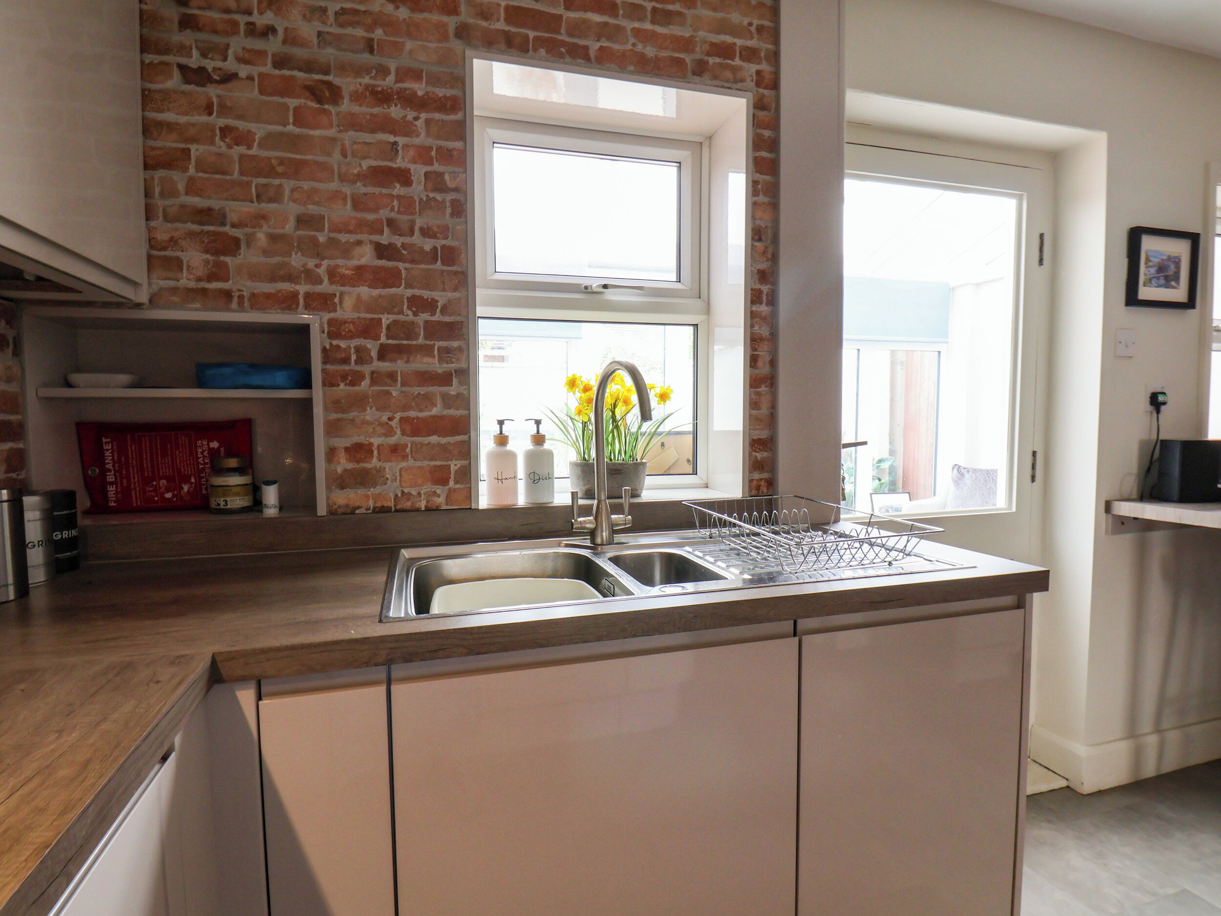 A kitchen with a brick wall, a window with flowers, a sink, and a shelf with kitchen items.