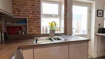 A kitchen with a brick wall, a window with flowers, a sink, and a shelf with kitchen items.
