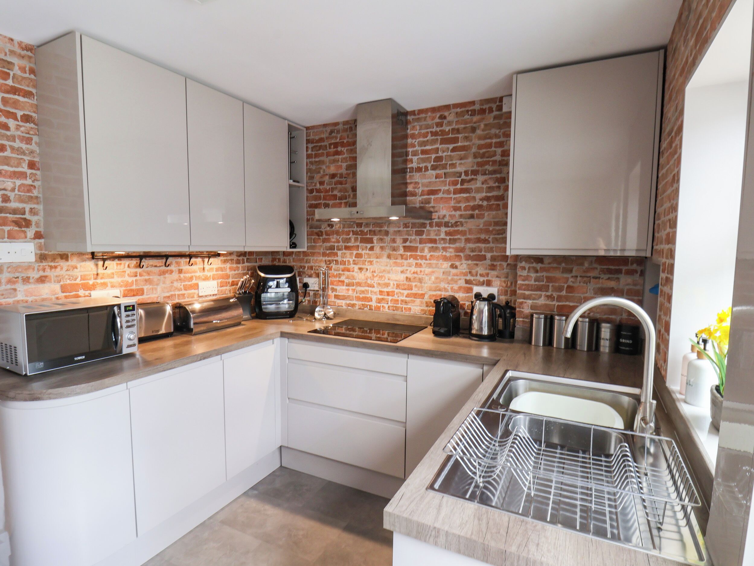 A compact kitchen with white cabinets, a brick wall, a stainless steel sink, and a dish rack.