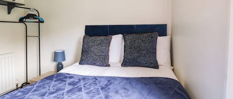 Een slaapkamer met een bed, hoofdbord, een aan de muur hangend schilderij, een radiator en een plank met spullen.