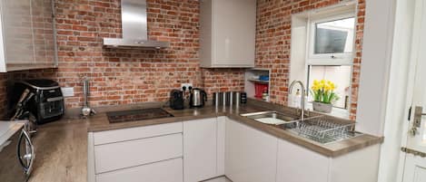 Een keuken met witte kastjes, een bakstenen muur, een roestvrijstalen afzuigkap, een spoelbak en een raam met bloemen.