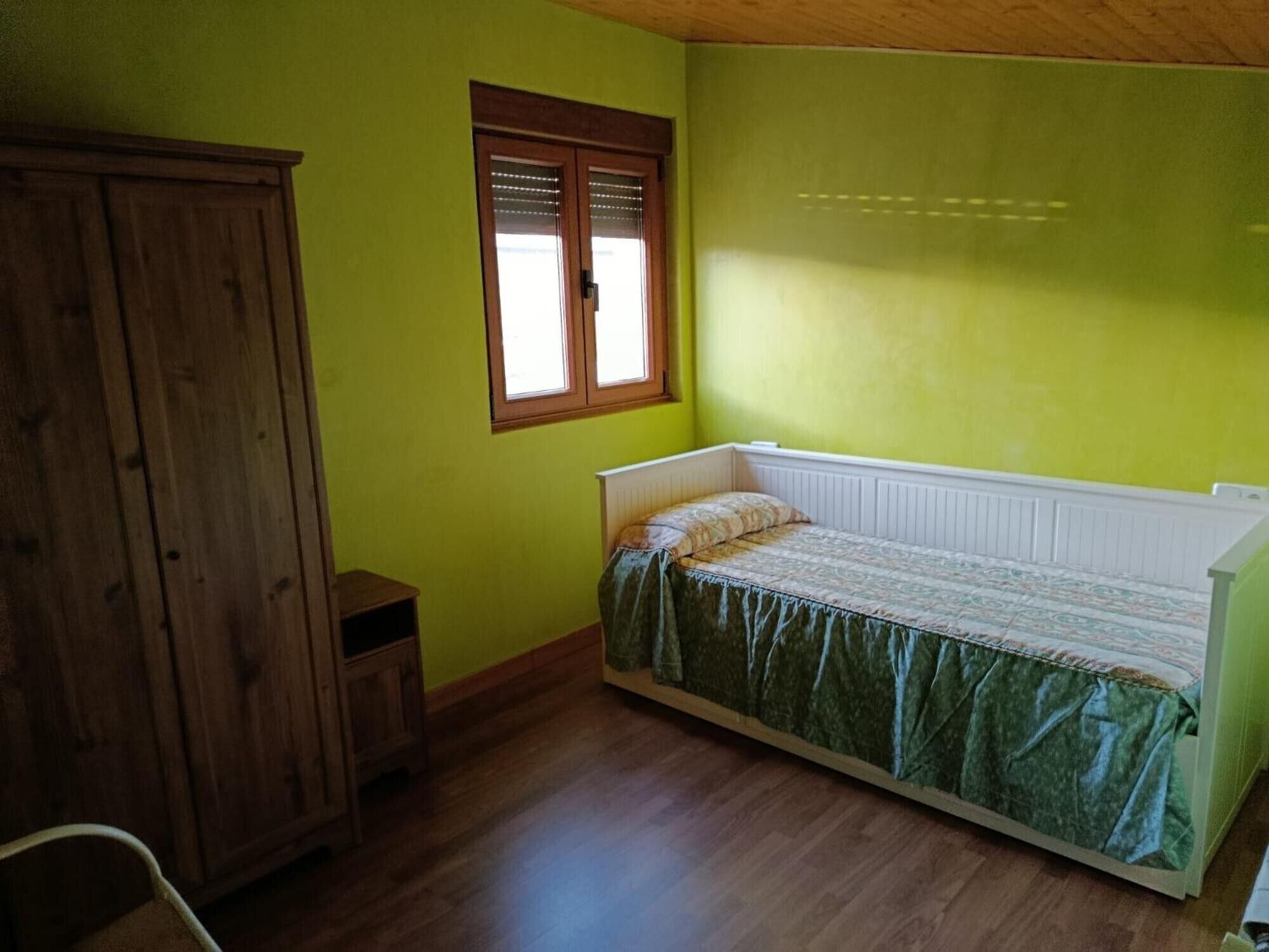 Une pièce avec une armoire en bois, un lit simple recouvert d’une courtepointe à motifs, une petite fenêtre avec des stores et un radiateur placé sous le lit.