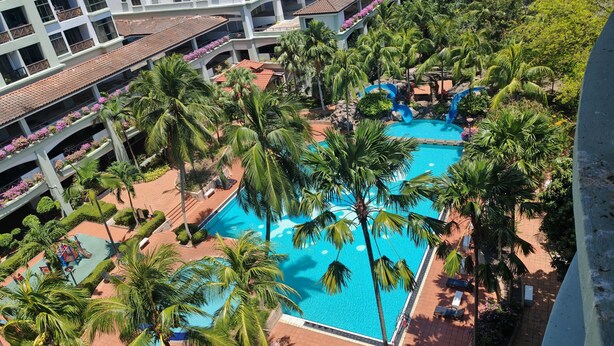 Ein mehrstöckiges Hotel mit einem zentralen Swimmingpool, umgeben von Palmen und einem Spielbereich für Kinder.