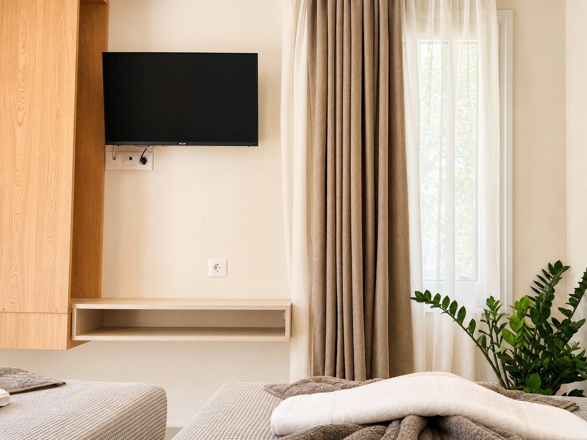 A hotel room with a flat-screen TV mounted on the wall, a window with curtains, a bedside plant, and a bed with a blanket.