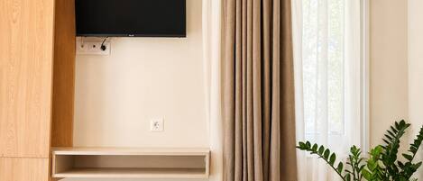 A hotel room with a flat-screen TV mounted on the wall, a window with curtains, a bedside plant, and a bed with a blanket.