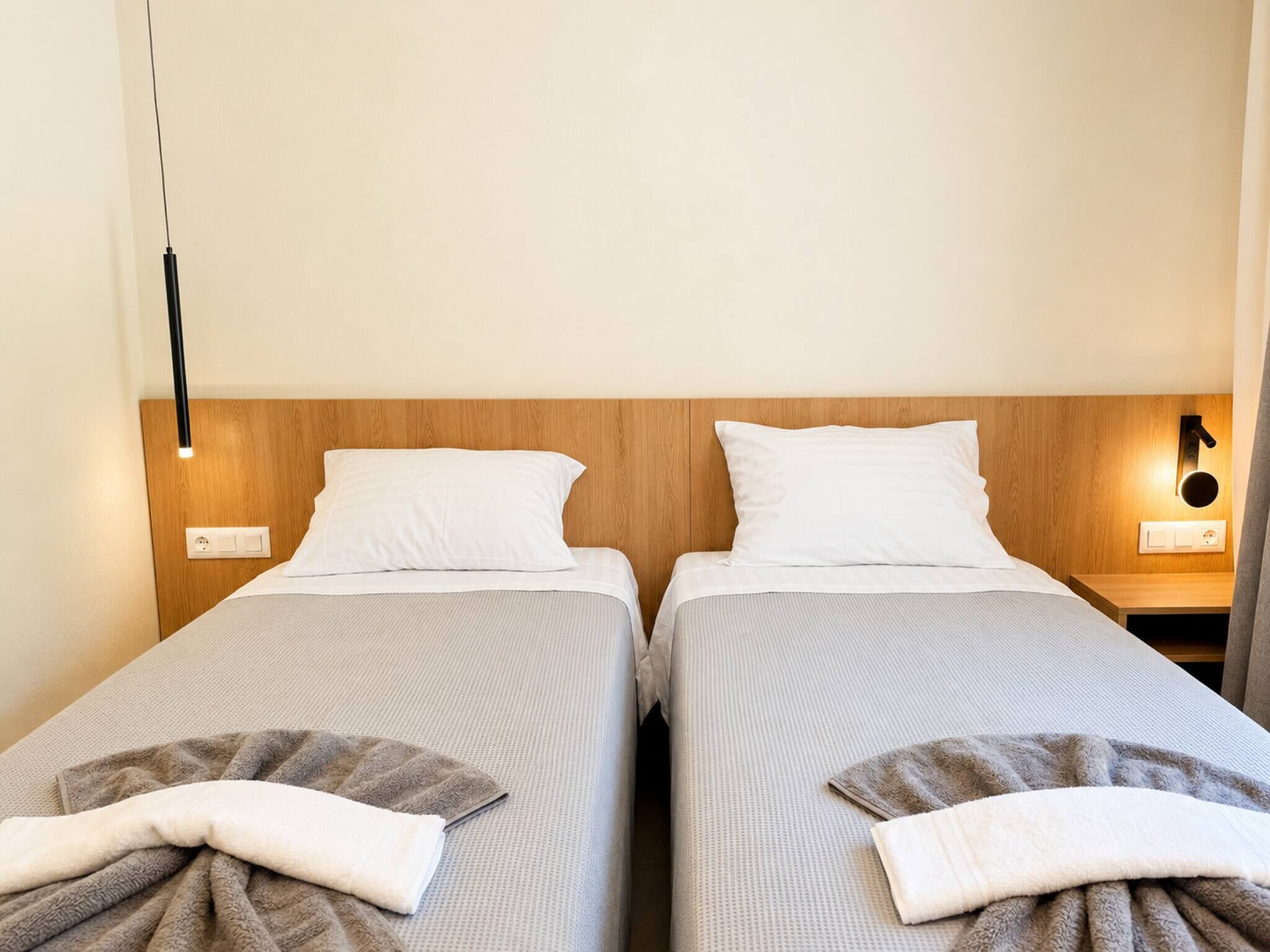 A hotel room with two single beds, wooden headboards, bedside tables with lamps, and a neutral color scheme.