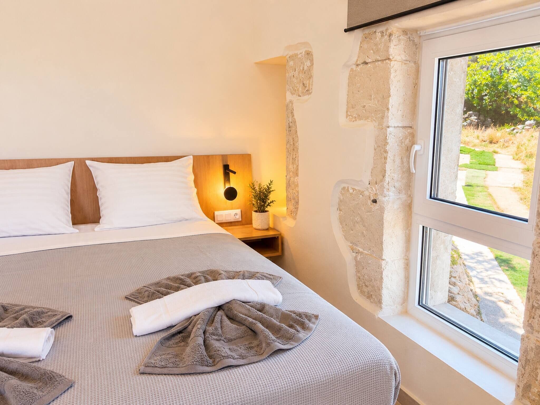 A bedroom with a bed, a bedside table with a lamp, a window with a view of greenery, and stone walls.