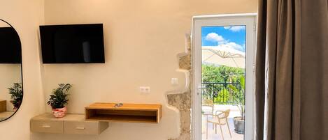 Ein Zimmer mit an der Wand befestigtem Fernseher, einem Holzbodenregal, einem Blumentopf und Blick auf einen Balkon mit Gartenmöbeln.