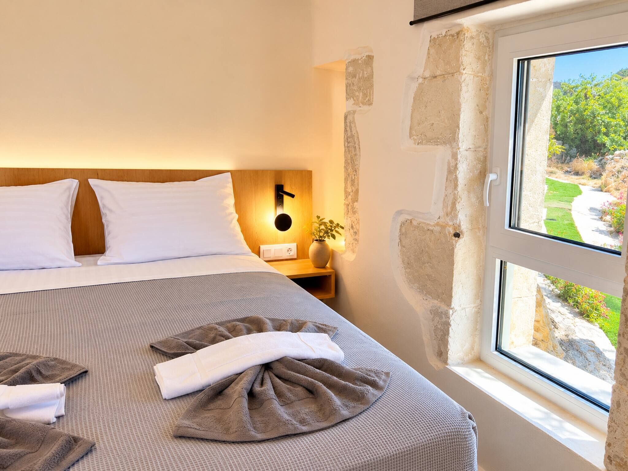 A neatly made bed with white pillows, a striped bedspread, and folded towels. There is a bedside table with a lamp and a vase with a plant. A stone wall and a window with a view of greenery are visible.