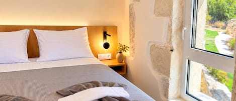 A neatly made bed with white pillows, a striped bedspread, and folded towels. There is a bedside table with a lamp and a vase with a plant. A stone wall and a window with a view of greenery are visible.