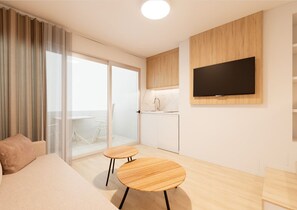 A modern hotel room with a sofa, two small wooden tables, a TV mounted on the wall, a kitchen area, and a sliding glass door leading to a balcony.