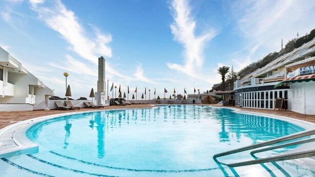 Ein Schwimmbecken mit blauem Himmel, Liegestühlen und einem Gebäude mit der Aufschrift 'Rest & Longevity'.