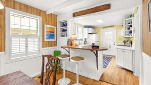 A kitchen with wooden cabinets, a bar area with stools, and a window with blinds. - Magical Coastal Cottage, Short Walk to Beach by Tybee Cottages (Tybee Island)